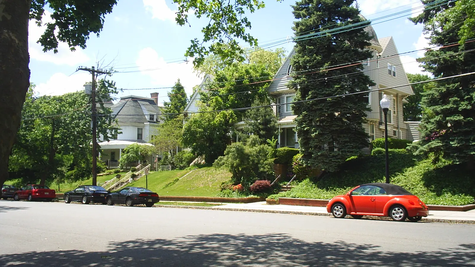 Tree-lined residential street in Perth Amboy, New Jersey, with large Victorian houses on a grassy rise and cars parked at the curb