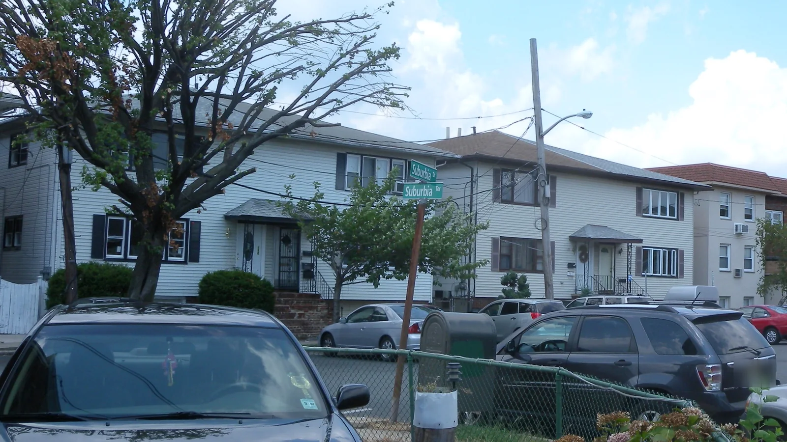Two-story attached houses with side-by-side front doors at Suburbia Drive and Suburbia Terrace in Jersey City, New Jersey, with cars parked along the street
