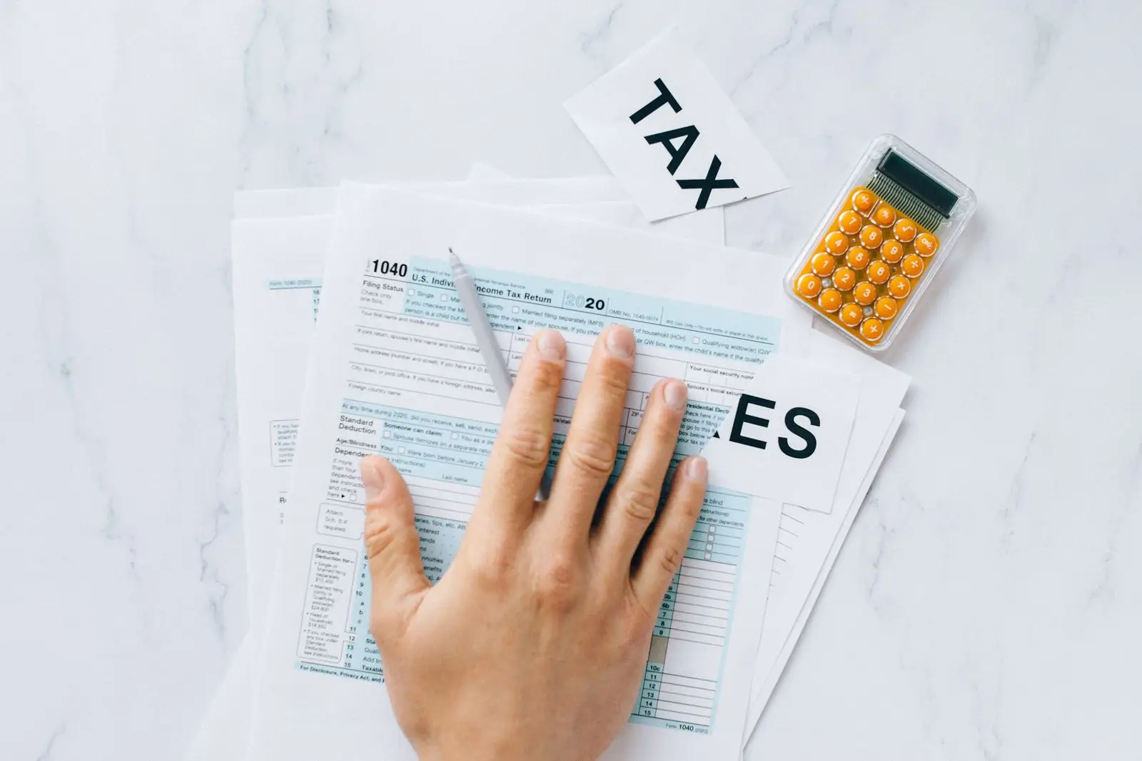A hand resting on a Form 1040 individual income tax return next to a pen and an orange calculator on a white marble desk