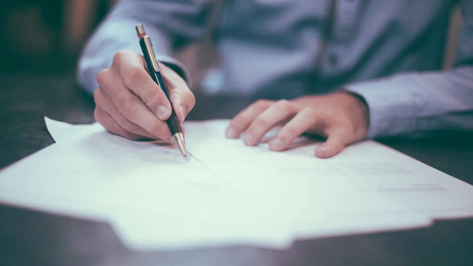A person in a light blue shirt holds a pen over a printed contract on a dark desk, about to sign