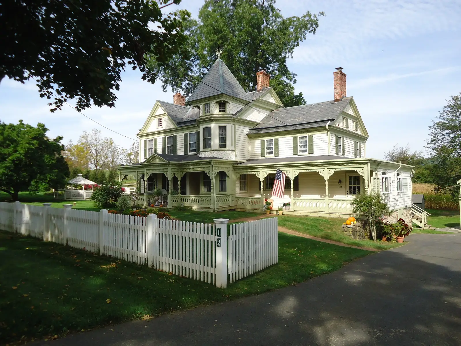 A cream Victorian house with a turret, green shutters and a wraparound porch behind a white picket fence and a front lawn in Oldwick, New Jersey