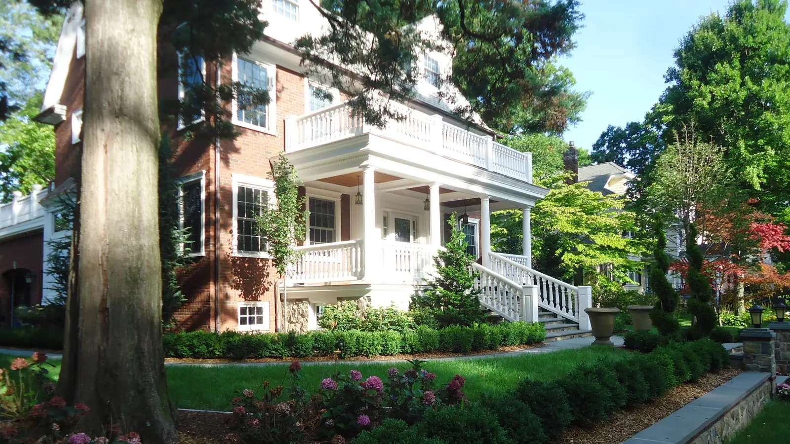 Brick colonial house with a white two-story porch, balustrade and front steps behind a landscaped lawn and stone wall in Summit, New Jersey