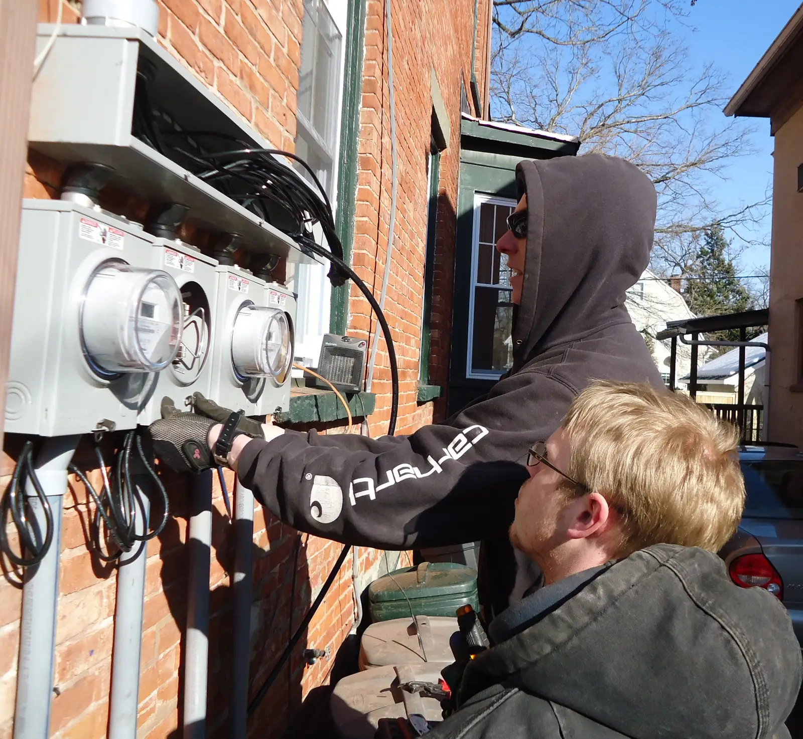Two electricians mount new meter panels on the brick wall of a multi-family house in New Jersey on a sunny winter day