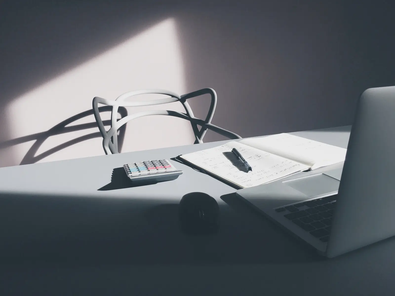 A desk with an open notebook and pen, a calculator and a laptop in afternoon light, the kind of setup used to total a year of bank deposits