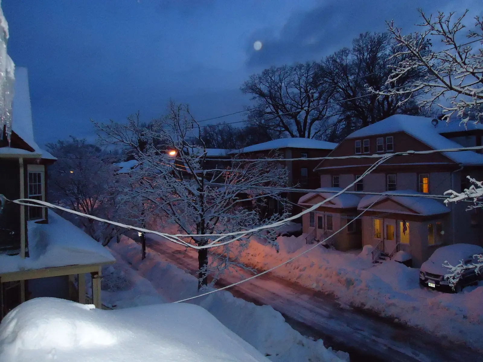 A snow-covered street of two-story houses with porches in Summit, New Jersey, at dawn with the moon still up
