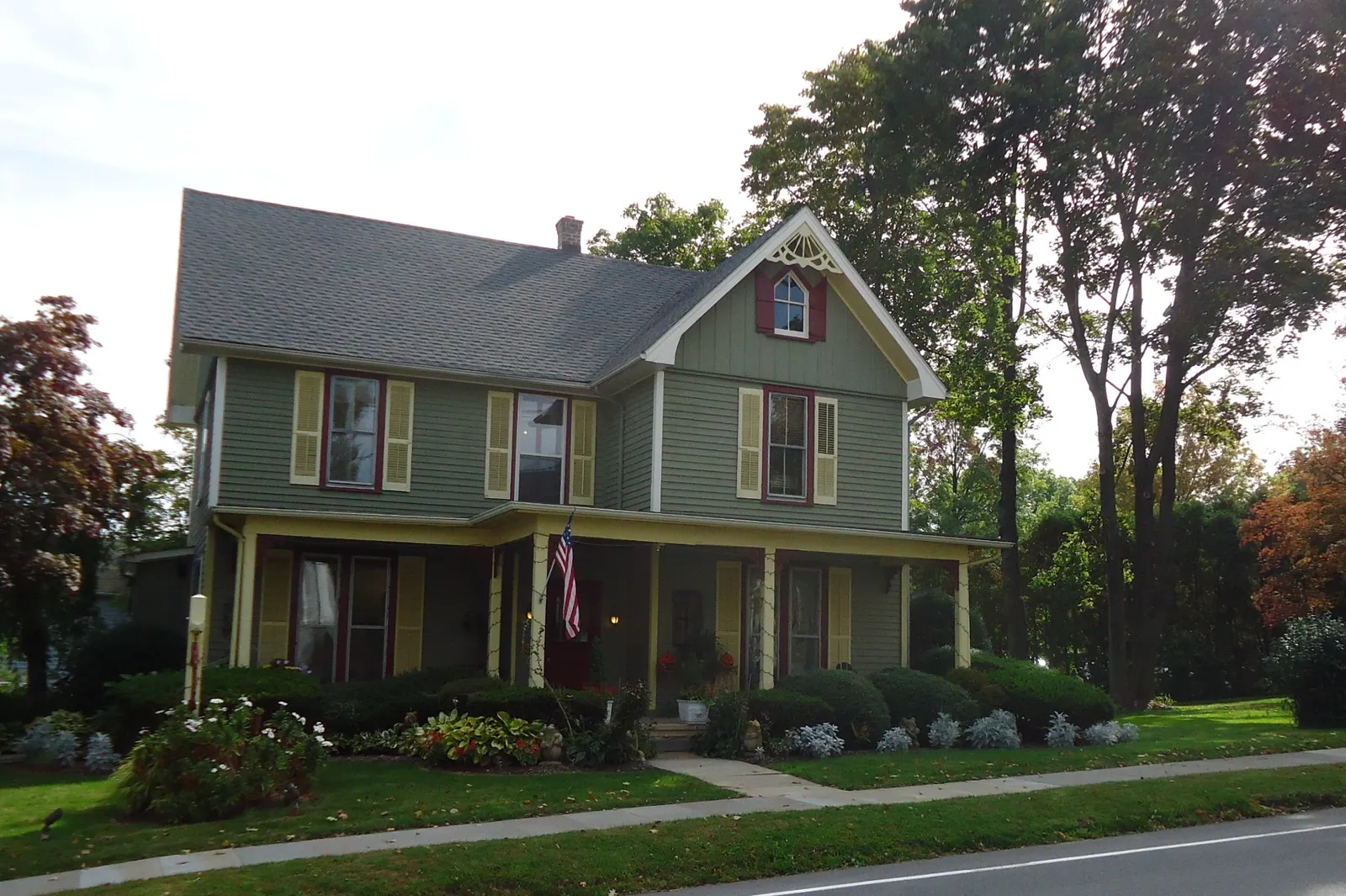 A two-story green house with yellow shutters, a front porch and an American flag behind a lawn and flower beds in Oldwick, New Jersey