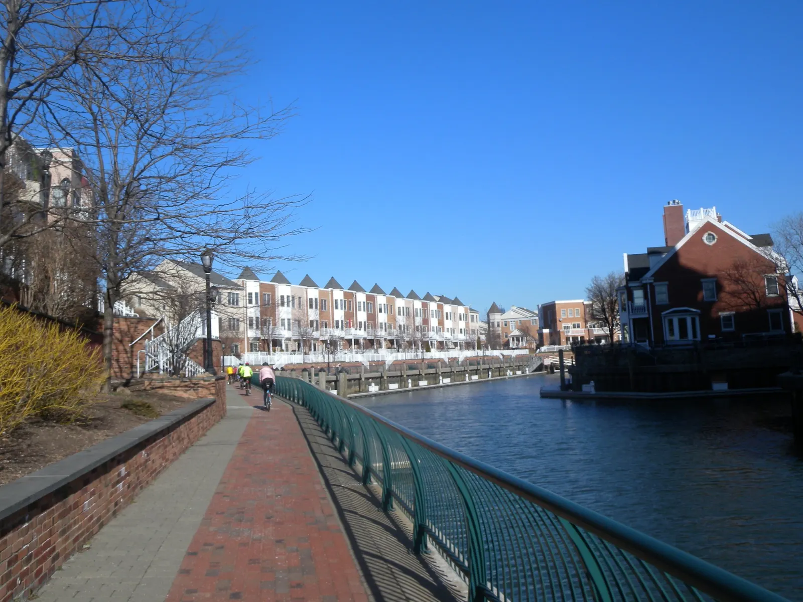 A row of three-story townhomes across a canal from a brick walkway with cyclists, under a clear blue sky at Port Liberté in Jersey City